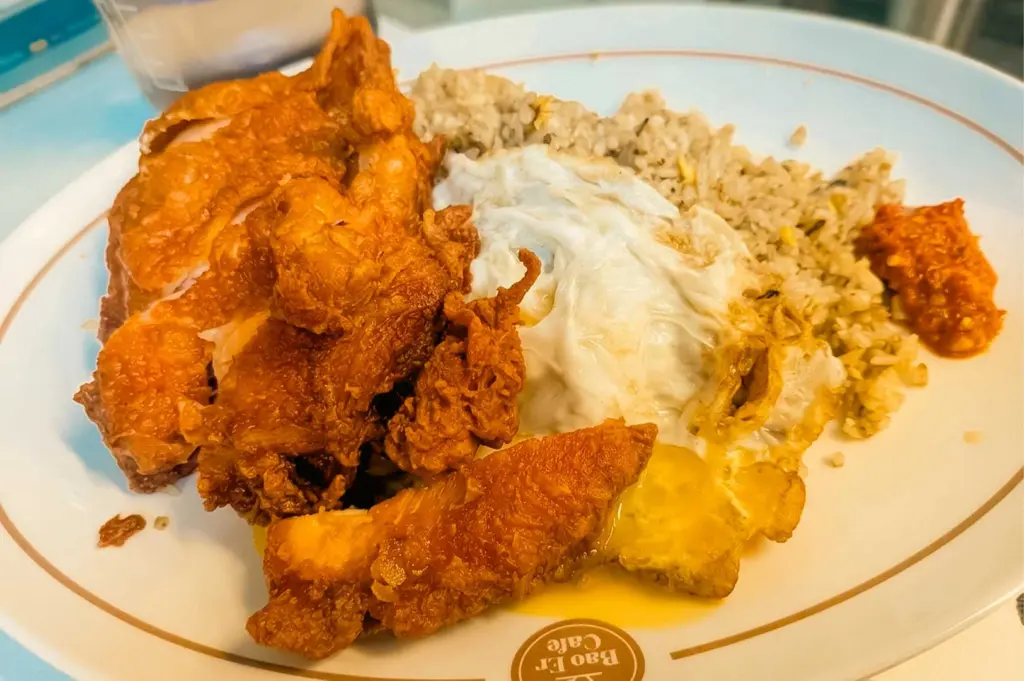 This close-up shot features a fried chicken cutlet rice dish served on a white ceramic plate bearing the "Bao Er Cafe" logo along the rim. The plate holds sliced, golden-brown deep-fried chicken with a crispy crust, a fried egg with a soft yolk resting in the center, a serving of seasoned rice, and a dollop of red sambal chili paste on the side.