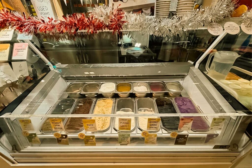 An illuminated glass freezer display case at `Hundred Acre Creamery` presents two rows of rectangular stainless steel tubs filled with colorful gelato and sorbet flavors, including yellow mango sorbet, coconut sorbet, and bright purple gelato. Small printed labels with handwritten-style typography mark each flavor along the front glass edge, while red and silver tinsel garland decorates the top ledge above stacked paper cups and water pitchers.