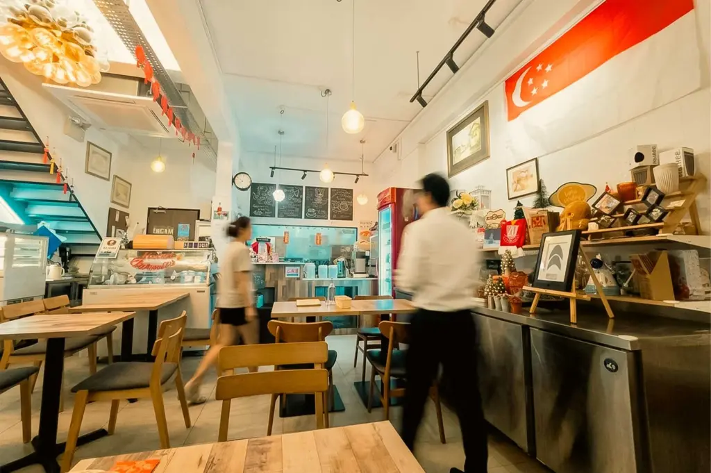 This wide-angle interior view captures the active dining room of Marie's Lapis Cafe, showing motion-blurred staff walking past wooden tables and chairs under hanging pendant lights. A dessert display case stands on the left near a blue staircase, while the right side features a long stainless-steel counter laden with shelf displays, framed artwork, and a Singapore flag mounted high on the white wall.