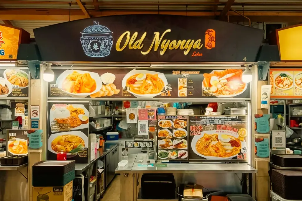 A straight-on, eye-level shot of the Old Nyonya hawker stall storefront. The dark blue overhead sign displays yellow script reading "Old Nyonya Laksa" alongside Chinese characters and an illustration of a classic blue-and-white porcelain pot. Below, illuminated menu boards show photos and pricing for signature dishes, including Laksa, Traditional Curry Chicken, and dry noodles, with additional menu posters mounted around the ordering counter.