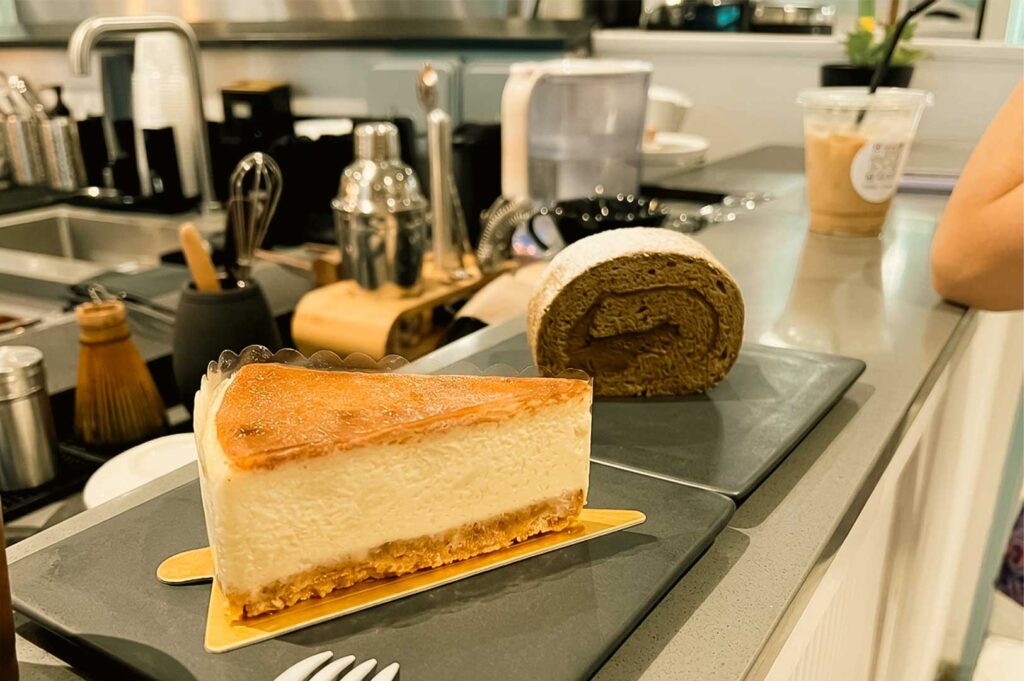A slice of burnt cheesecake resting on a gold cardboard base sits in the foreground on a slate serving plate, accompanied by a dark green hojicha roll cake dusted with powdered sugar. The desserts are displayed on a light gray cafe counter, with tea whisks, metal shakers, barista tools, and an iced drink visible in the background.
