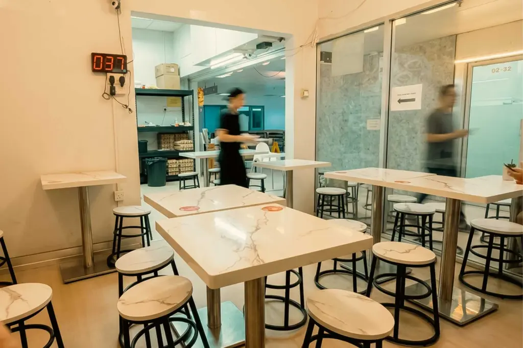 This interior shot showcases the indoor seating area of Bao Er Cafe, featuring white marble-patterned tables paired with round wooden stools on black metal frames. Warm overhead lighting illuminates the casual space, where glass partition walls line the right side and a red digital queue display box numbered "037" hangs on the back left wall above an open doorway leading to the kitchen and storage area.