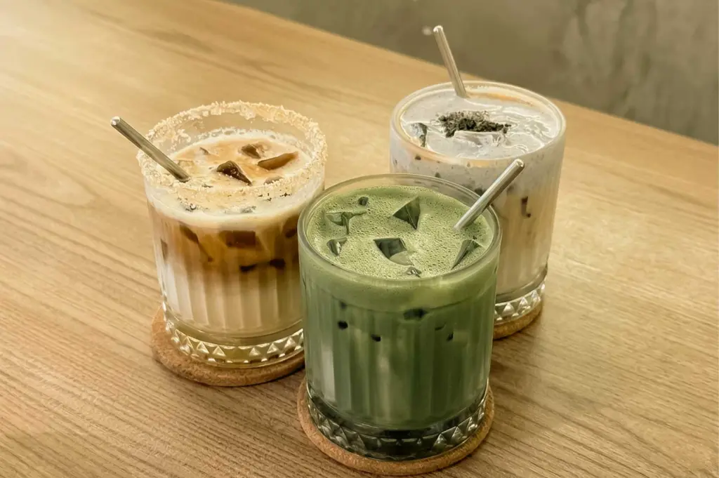 Three iced specialty beverages including an iced matcha latte, a sugar-rimmed coffee latte, and a black sesame drink with metal straws served on cork coasters at September Coffee.