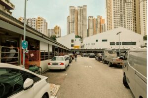A ground-level view of an outdoor parking lot situated between low-rise hawker stalls and residential buildings. Cars and a commercial pickup truck are parked along both sides of the narrow asphalt lane, while a solitary pedestrian walks down the center, framed by tall, modern high-rise apartment blocks towering in the background under a pale, overcast sky.