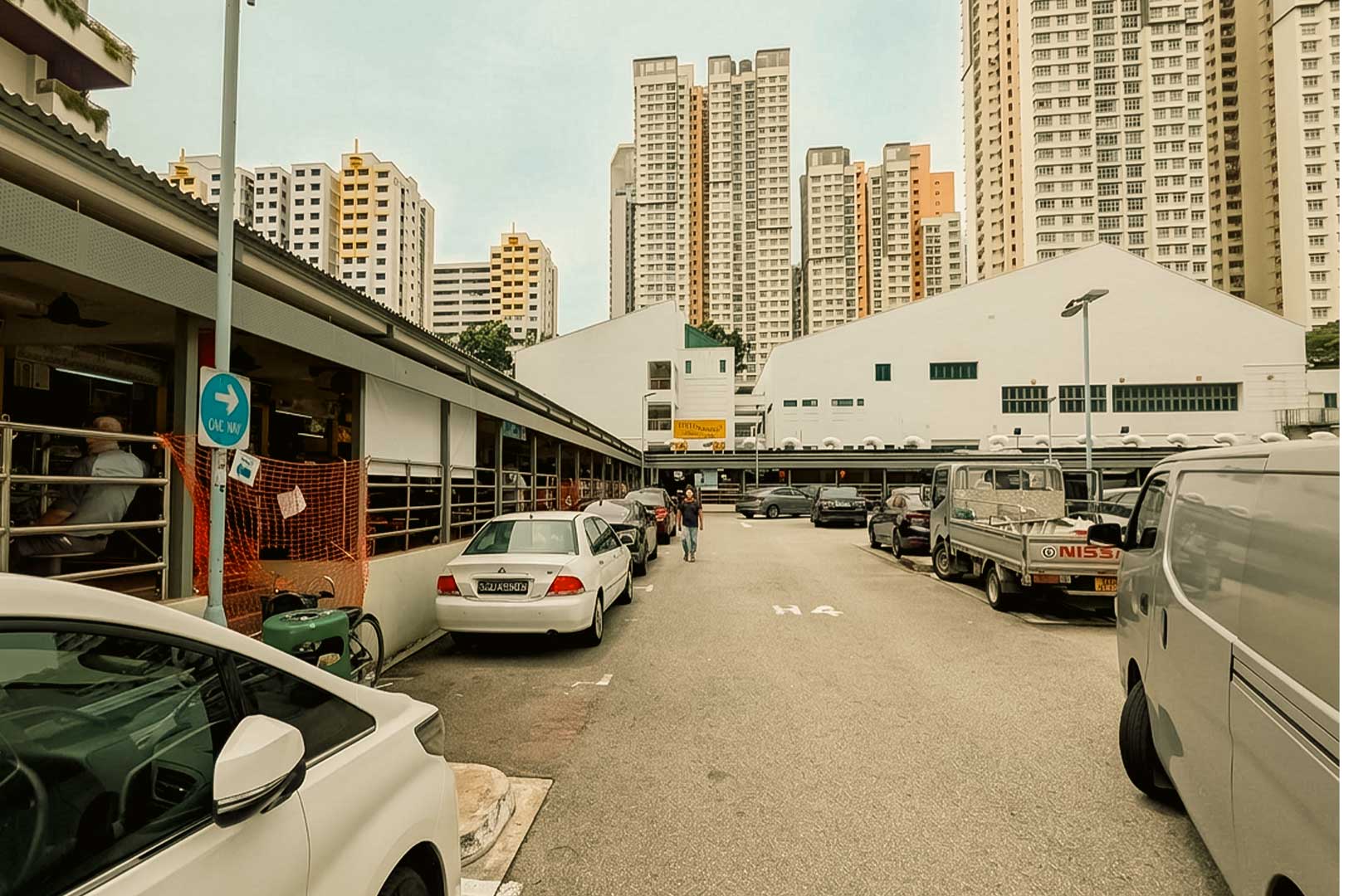 A ground-level view of an outdoor parking lot situated between low-rise hawker stalls and residential buildings. Cars and a commercial pickup truck are parked along both sides of the narrow asphalt lane, while a solitary pedestrian walks down the center, framed by tall, modern high-rise apartment blocks towering in the background under a pale, overcast sky.