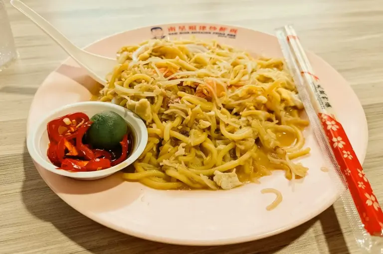 A plate of Singapore-style Hokkien fried prawn mee combining yellow thick noodles and thin rice vermicelli in light savory stock, served on a light pink oval plate alongside a plastic spoon, packaged disposable chopsticks, and a small white dish with sliced red chili and a calamansi lime half.