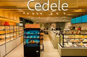 The storefront of a Cedele Bakery Kitchen branch featuring a prominent white illuminated "Cedele" sign overhead, a central "Fresh To Go" refrigerated display unit, bread shelves along the left wall, a service counter, and a glass cake showcase filled with whole cakes and pastries on the right.