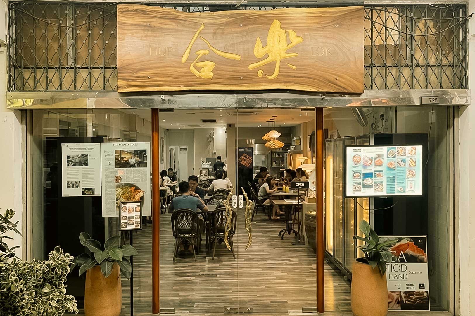 A wide, straight-on shot of the entrance to Hup Lok restaurant, featuring a large, natural-edge wooden sign above the glass doors displaying Chinese characters and the English name. The glass facade is decorated with potted plants, news article clippings, and an illuminated digital menu board, offering a full view into the brightly lit dining area where several customers sit at wooden tables.