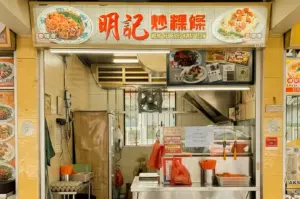 The front view of the Meng Kee Fried Kway Teow hawker stall (unit 01.67), featuring a large overhead banner with red Chinese typography and bilingual dish photography, a stainless steel order counter with plastic cutlery containers, and a compact kitchen interior complete with an exhaust hood, cooling fan, and framed media features.