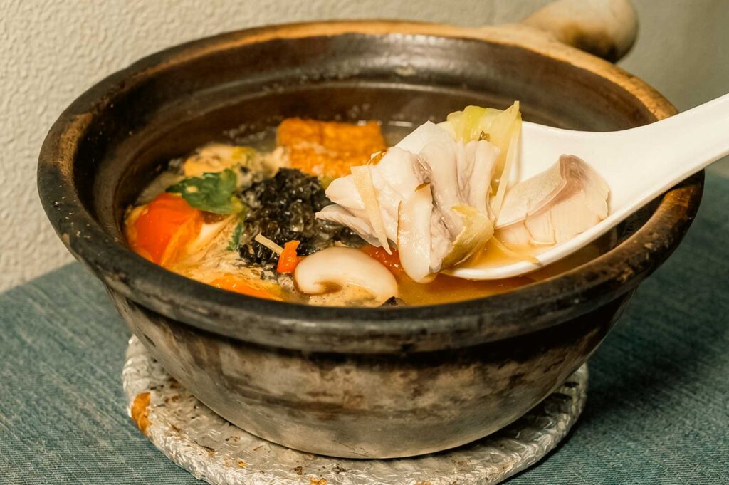 A claypot of hot fish soup from Hup Lok Fish Soup, featuring a white porcelain spoon scooping tender slices of fresh white fish and cabbage from a rich, savory broth filled with tofu, seaweed, tomatoes, and sliced mushrooms.