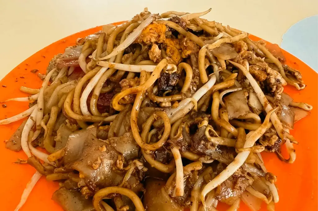 A generous plate of fried kway teow from Meng Kee Fried Kway Teow, served on a bright orange plastic dish and filled with stir-fried flat rice noodles, yellow egg noodles, crunchy bean sprouts, scrambled egg, cockles, and Chinese sausage coated in a glossy dark soy sauce glaze.