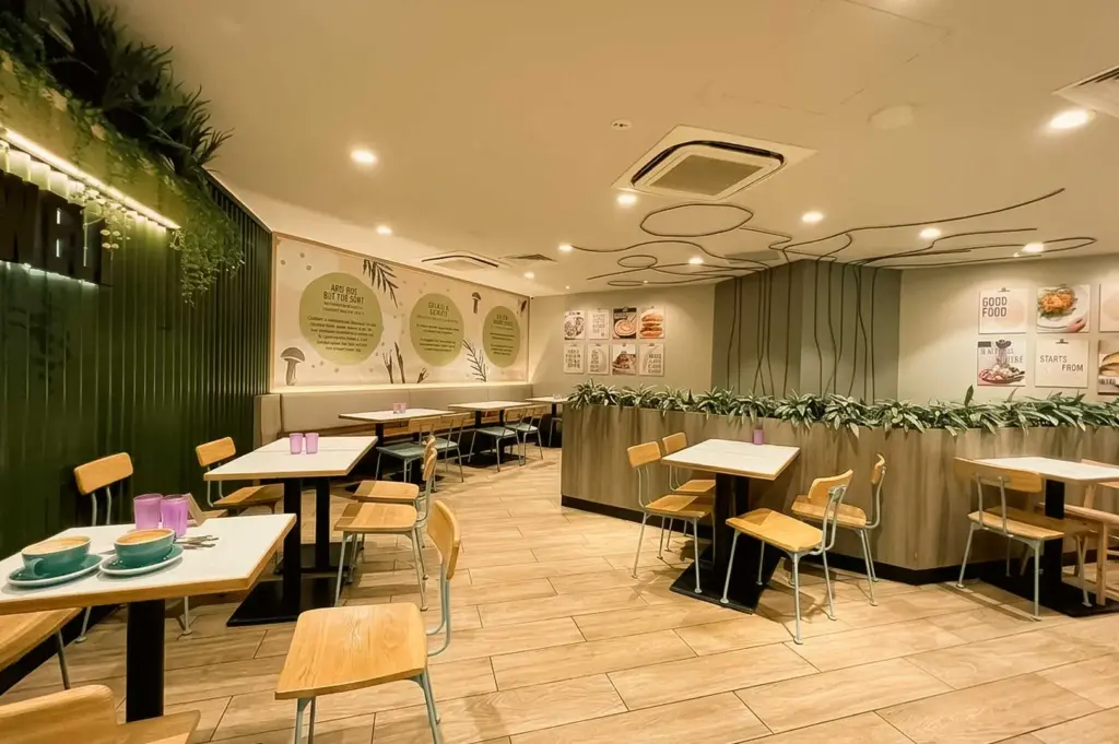 The warm and modern dining area of Cedele Bakery Kitchen, featuring light wood flooring, white tables with wooden chairs, green accent walls adorned with artificial foliage, and informative wall graphic displays.