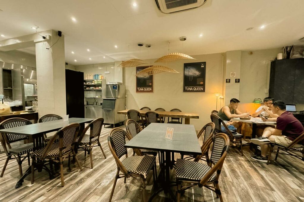 A wide-angle view of a brightly lit, modern restaurant interior featuring gray wood-look flooring, recessed ceiling spotlights, and woven cone-shaped pendant lights. Dark dining tables with checkered woven-style chairs are arranged throughout the space, while a group of patrons dines together in the background near walls adorned with framed "Tuna Queen" posters.