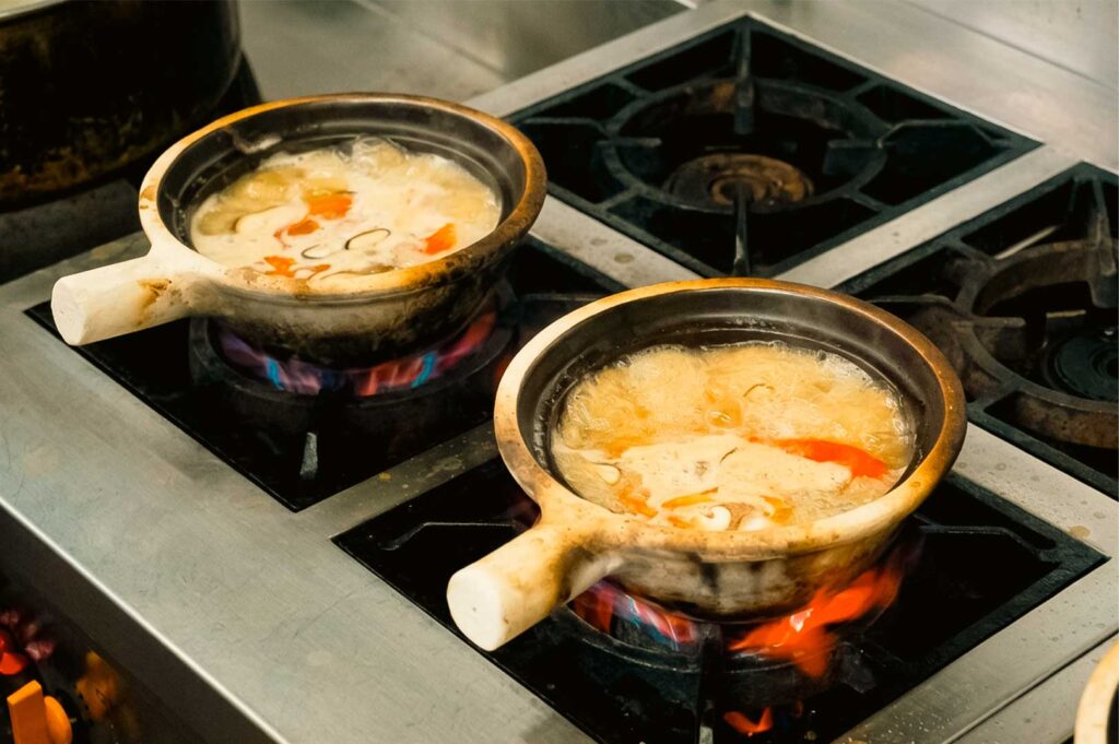 Two earthen claypots with light-colored handles simmering over open gas flames on a stainless steel commercial stovetop at Hup Lok Fish Soup, filled with bubbling, golden fish broth containing tomatoes, cabbage, and seafood.