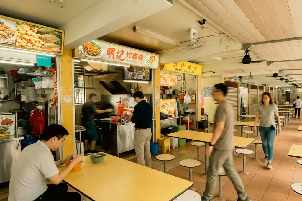 A wide shot of a bustling hawker center alleyway centered on the Meng Kee Fried Kway Teow stall, showing customers walking past, a diner seated at a yellow laminate table enjoying a meal in the foreground, and a customer waiting at the counter while a cook prepares food inside the stall.